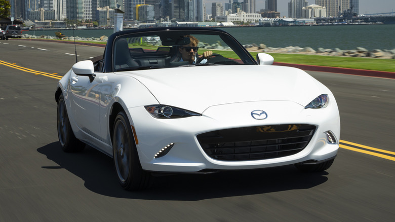 A Mazda MX-5 Miata driving on a road with a body of water and a city skyline in the background