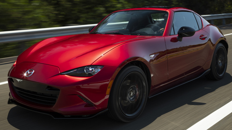 A Mazda MX-5 driving on a paved road with white lane markings and a metal guardrail