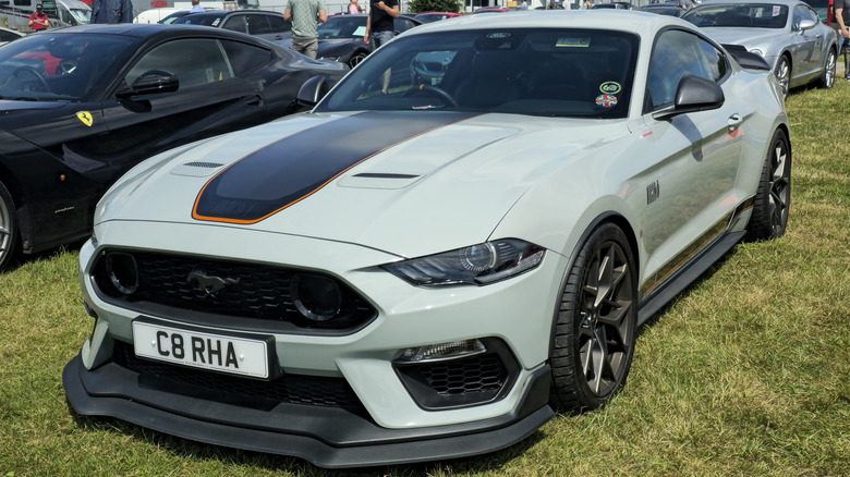 3/4 view of a 2021 Ford Mustang Mach 1 parked on grass with other cars in the background.