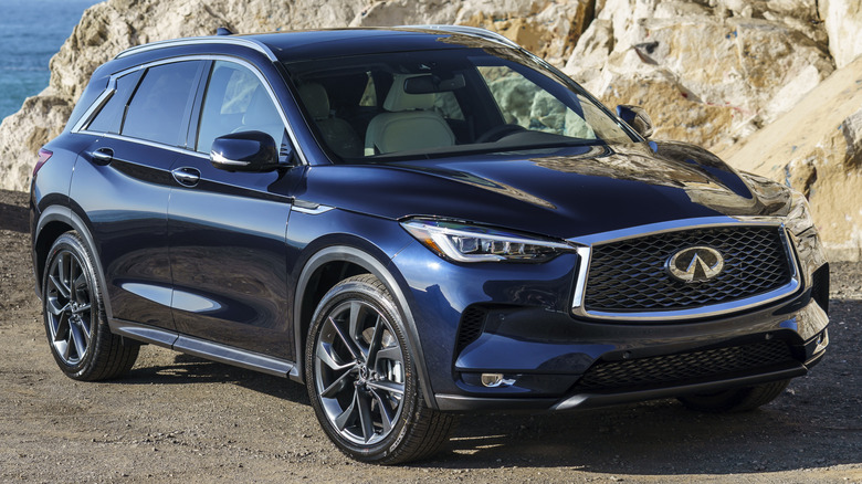An Infiniti QX50 in Hermosa Blue, parked in front of water and a rocky hill.