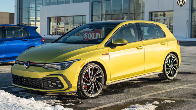 A yellow Volkswagen Golf GTI parked in front of a VW dealership in the snow.