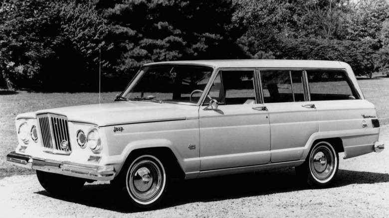 A black-and-white image of a 1963 Jeep Wagoneer parked in front of grass and trees
