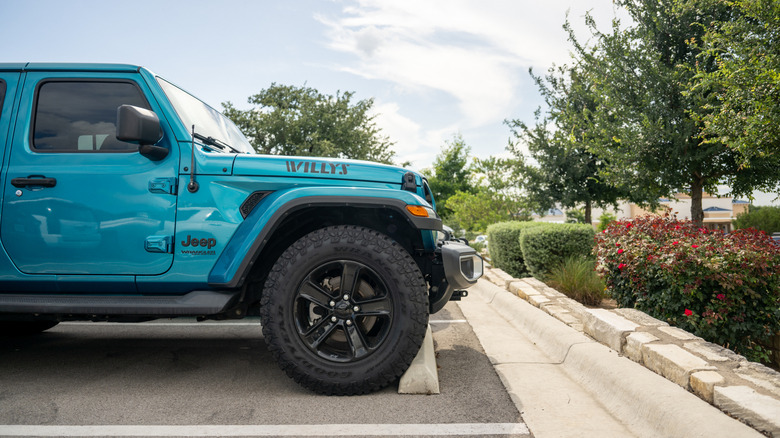 A front-side profile view of a light blue Jeep Wrangler parked in front of hedges and greenery.