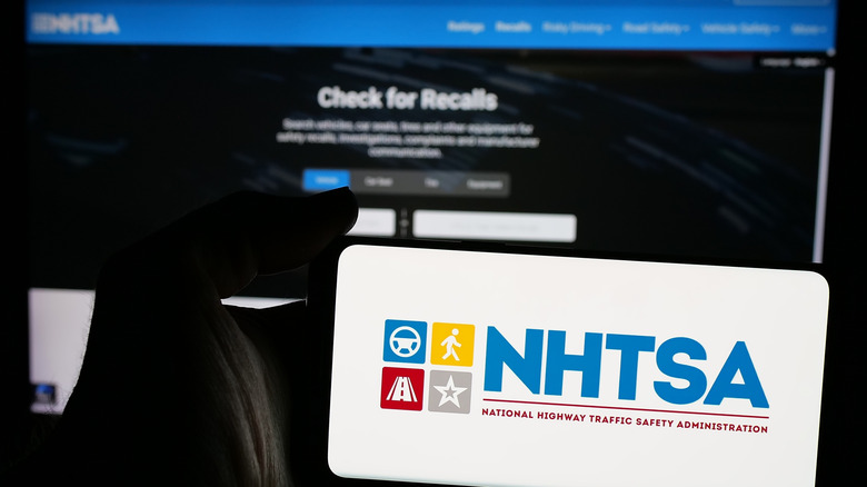A smartphone with an on-screen NHTSA logo in front of a monitor displaying the NHTSA recall website.