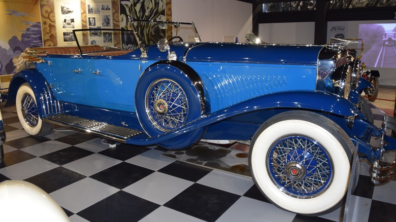 A blue 1931 Duesenberg Model J parked on a checkerboard floor at a car museum