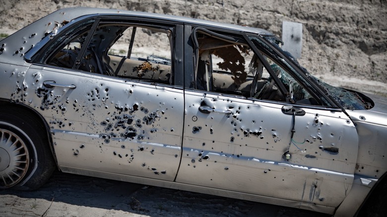 A junk car with bullet holes in a rocky area