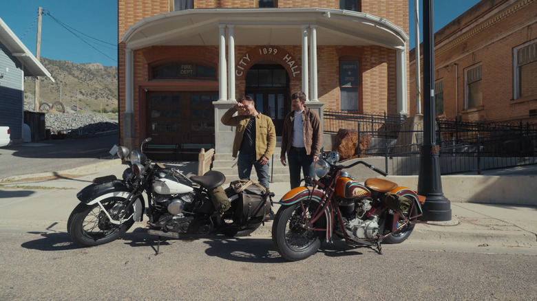 Two men stand behind two vintage motorcycles