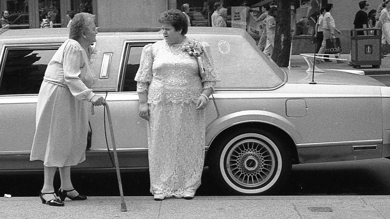 A black and white photo of two women standing next to a limousine with a boomerang antenna on the trunk.