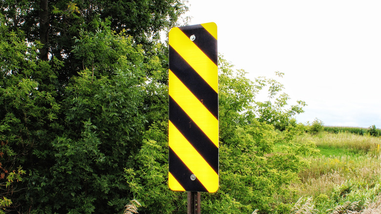 An OM-3L black and yellow diagonal stripe road sign with bushes in the background.
