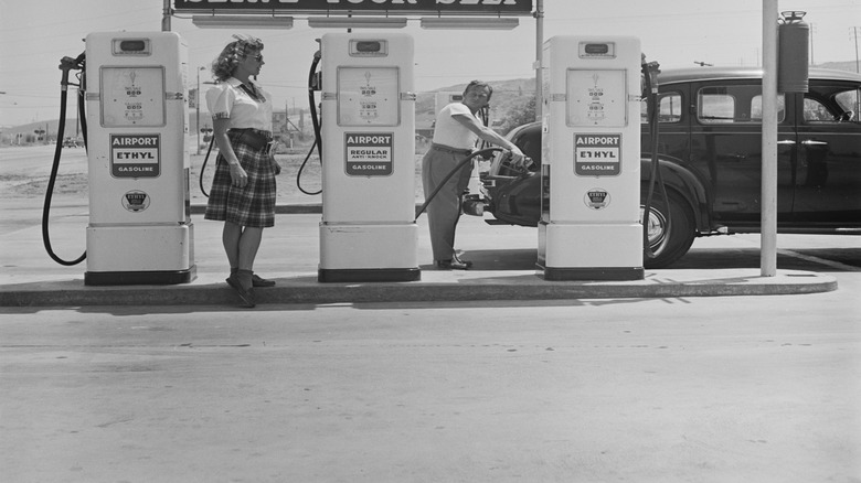 A woman in a plaid skirt, with a change dispenser around her waist, stands between fuel pumps as a man uses a pump to refuel his car at a self-service gas station, United States, circa 1945.