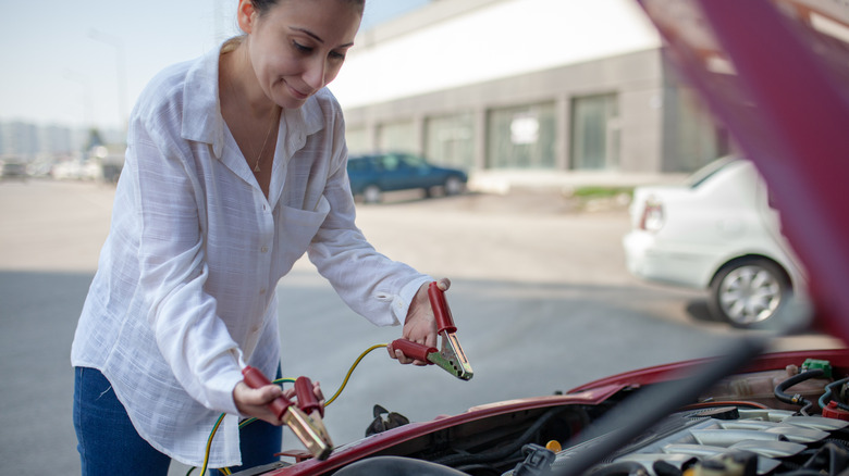 A person attaches alligator clips to a car's battery in a relatively empty parking lot