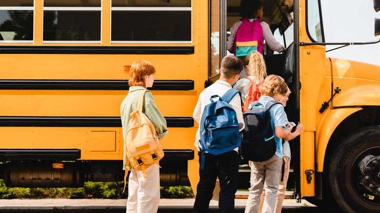 A small gaggle of children clad in backpacks wait in line to board a large yellow school bus.