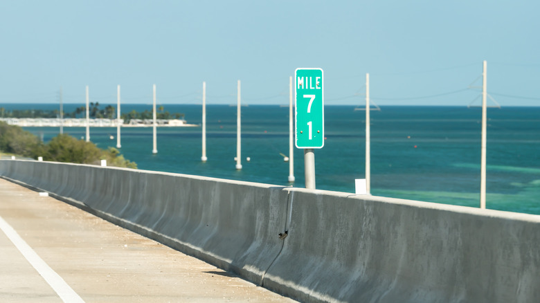 A green mile-71 mile marker, posted on a bridge with the ocean in the background.