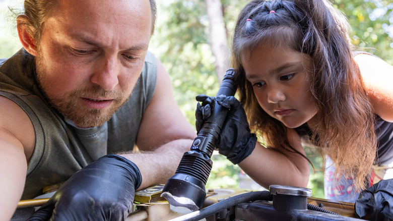 A young girl holding a flashlight over the engine of a car, as an adult teaches her how to fix it