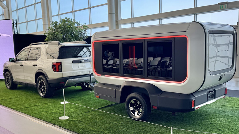 A rear three quarters shot of the Base Station trailer hitched up to a white Passport TrailSport parked on astroturf in a building in front of frosted windows