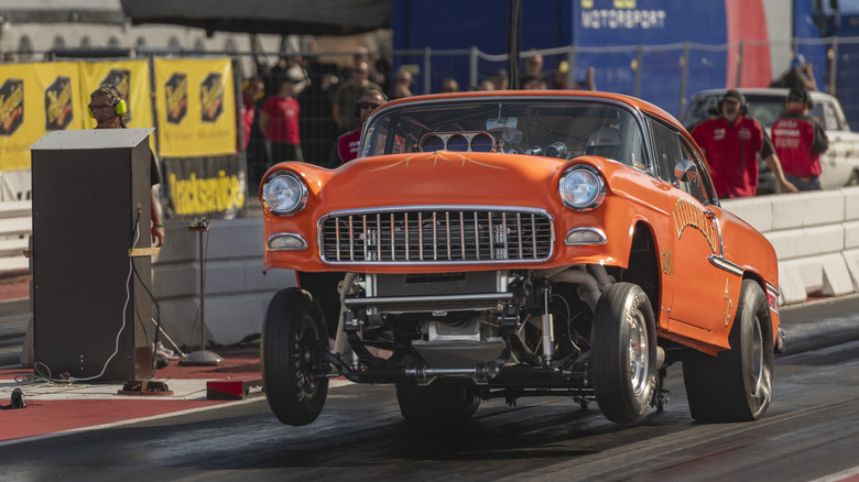 Orange 1950s Chevrolet Gasser hot rod seen in left front three-quarter view popping a wheelie on a drag strip
