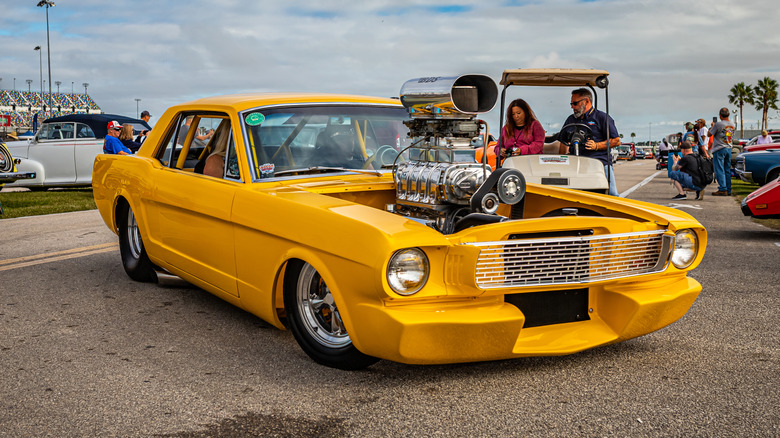 Yellow 1965 Ford Mustang pro street style hot rod with a towering motor, roll cage, and fat rear tires, seen in right front three-quarter view