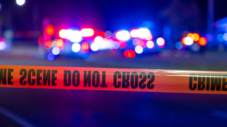 Police tape reading "crime scene do not cross" stretches across blurred cop lights in a nighttime scene.