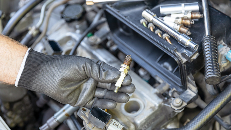 A hand in a black glove holding a spark plug above a four-cylinder engine.