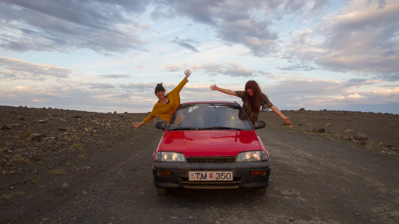 two girls in a red Suzuki Swift under a cloudy sky in a desert