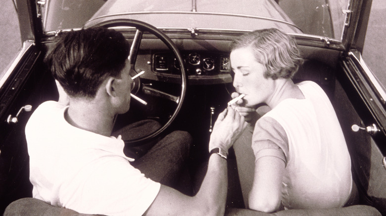 a boy lights a girl's cigarette in an open top car. black and white photograph