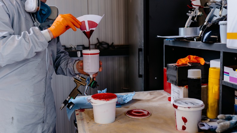 Red automotive paint being poured into a vertical-feed spray gun by a painter in protective gear.