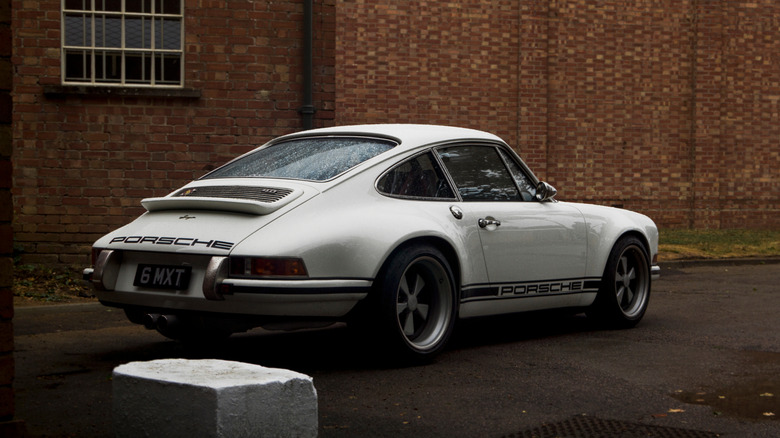 A white Singer Reimagined Porsche 911 parked near a red brick building.