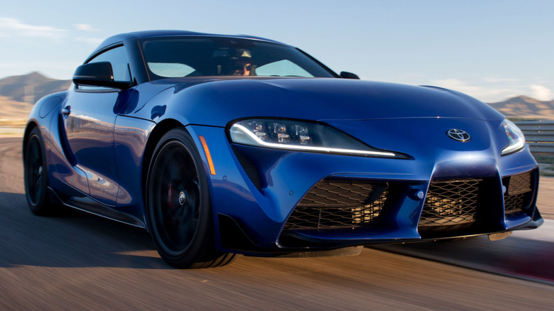 A blue 2025 Toyota GR Supra driving on a track in the desert with mountains behind it.