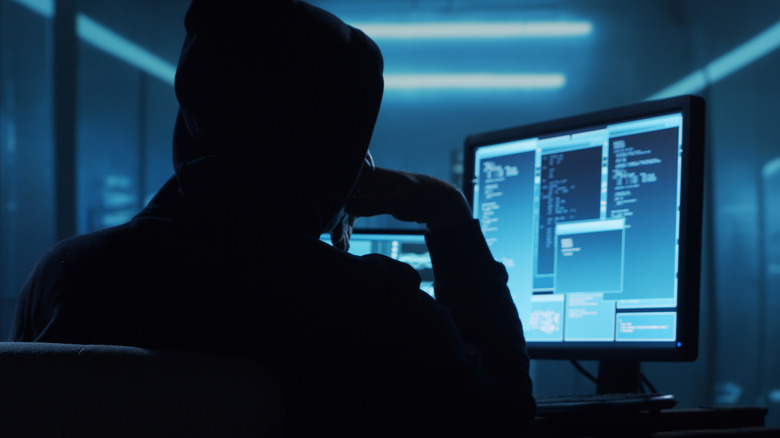 A stock image of a hooded hacker in a dark room sitting in front of multiple computer monitors