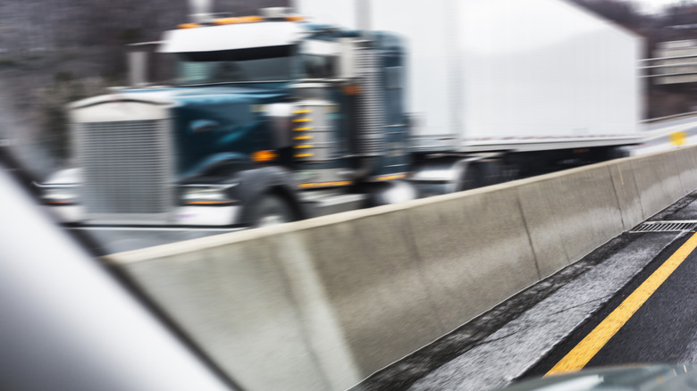 A tractor trailer speeding by in the opposite lane of a roadway divided by a single slope concrete highway barrier.