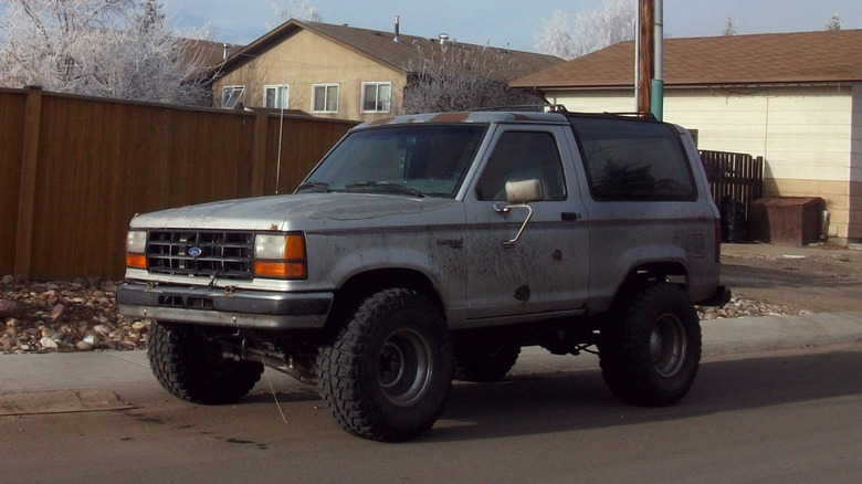 An old and rusted silver Ford Bronco II parked next to a sidewalk.