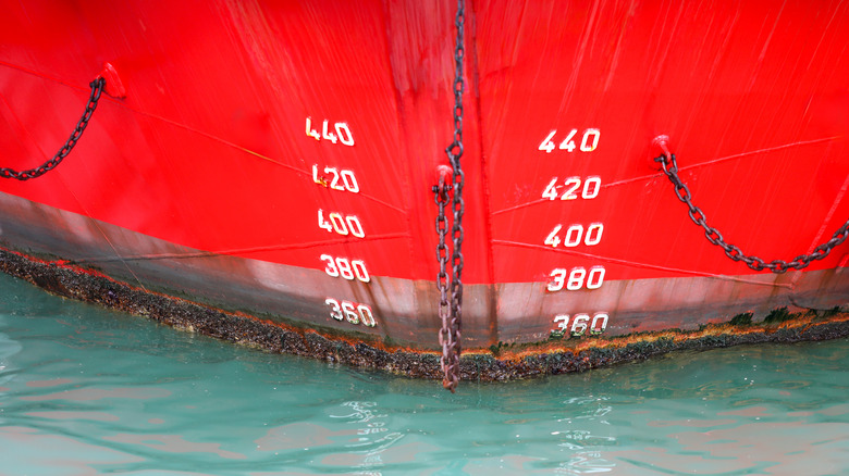 Red ship's bow with white draft marks and an anchor chain emerging from the water, displaying the waterline where barnacles cling to the aged hull, signifying ocean travel and freight
