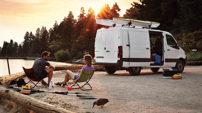 A Mercedes Sprinter camper van parked on a lakeside with a couple enjoying the sunset