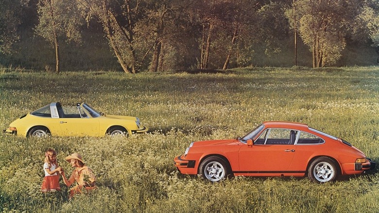 A yellow Porsche 911 Targa and red 911 Targa coupé from 1976 in a field with high grass and a woman and child in the foreground