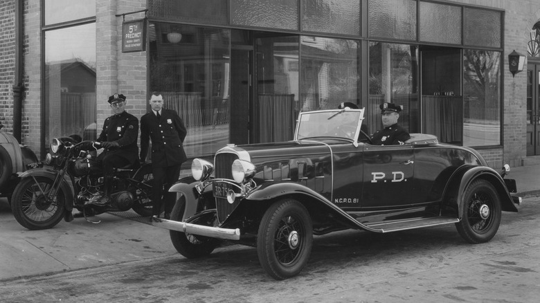 A black-and-white photograph of officers in a police car and on motorcycle outside a police station in 1929.