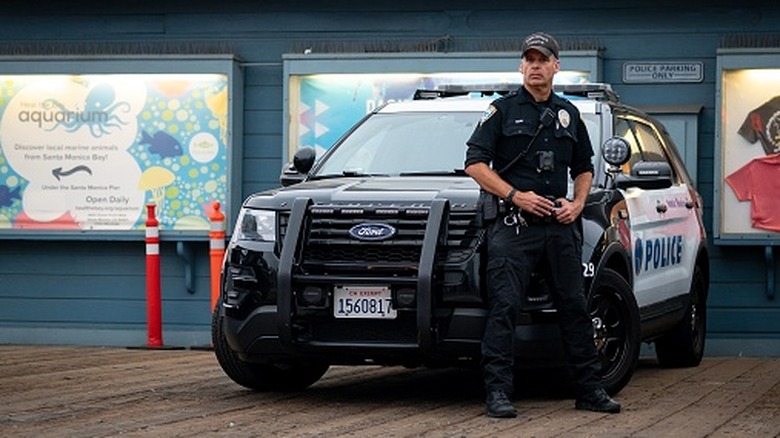 A policeman with a serious expression next to his Ford Police Interceptor SUV, keeping watch on the Santa Monica pier.