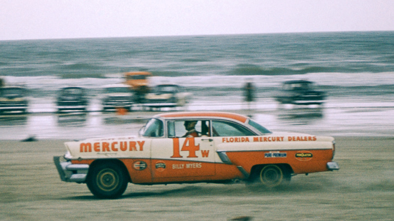 A NASCAR race in 1956. An organe and cream car sponsored by Florida Mercury dealers speeds down a beach with other cars in the background and the ocean behind them.