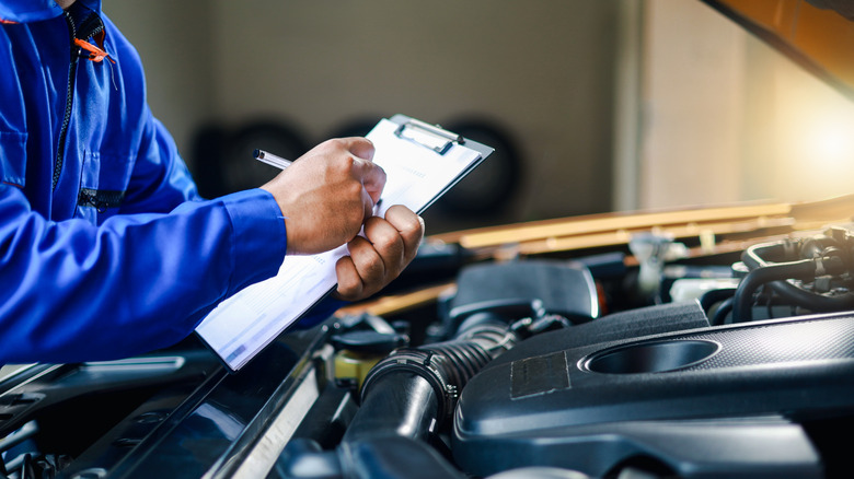 A person going through a checklist in front of a car with an open hood.
