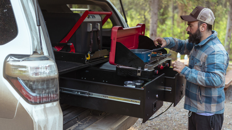 A bearded man cooking on a two-burner gas stove while camping in his car.