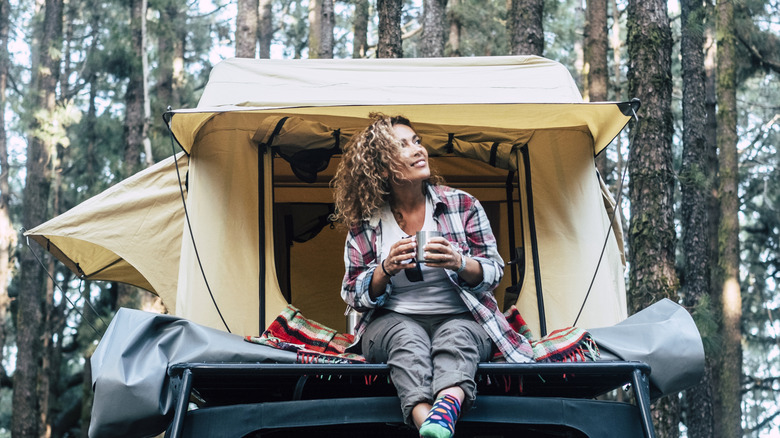 A woman drinking a cup of coffee while camping in her car in the woods.