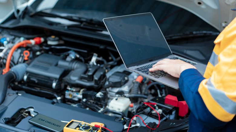 A technician wearing a safety shirt and holding a laptop, using diagnostic tools to assess a vehicle.