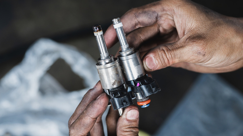A technician holding an old and a new fuel injector beside each other with dirty hands.