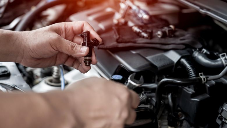 A technician replacing a black fuel injector under the hood of a vehicle with no gloves on.