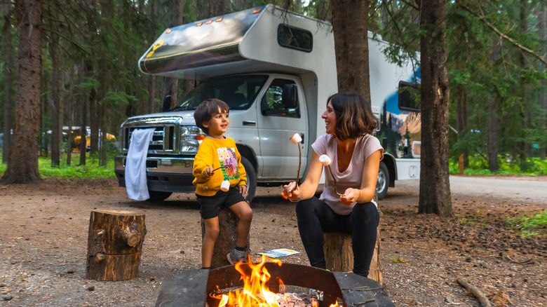 A mother and son roasting marshmallows over a campfire in front of an RV parked in a forest