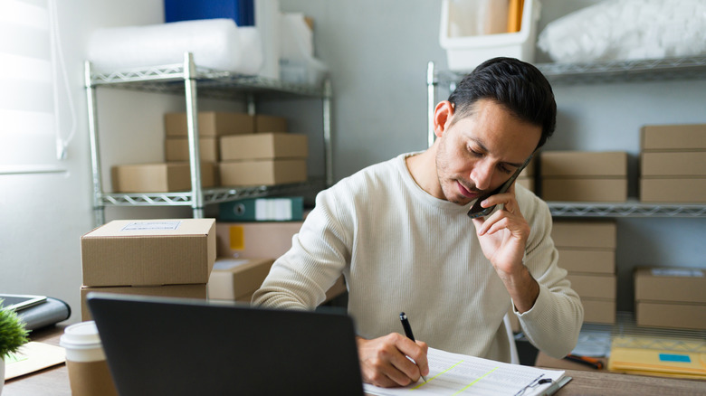 A person talking on the phone and taking notes with boxes on shelves in the background