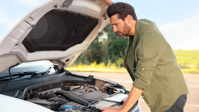 A bearded man wearing a green shirt examining a car's engine on a sunny day.
