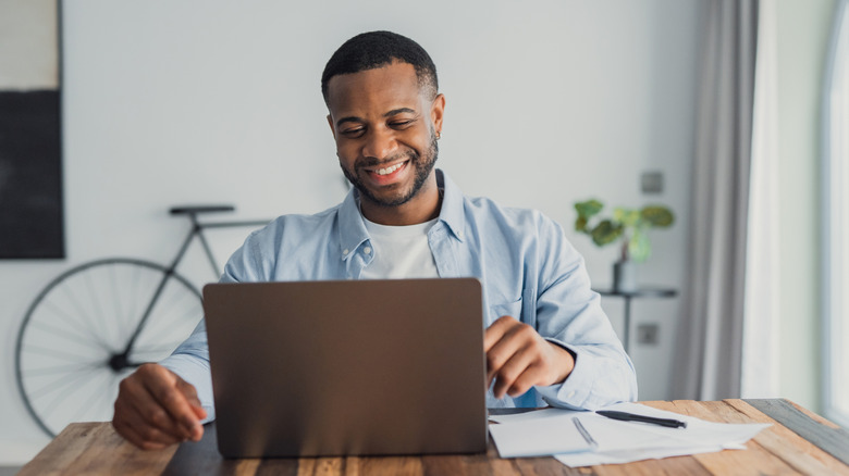 A man smiles while looking at his computer with papers nearby.