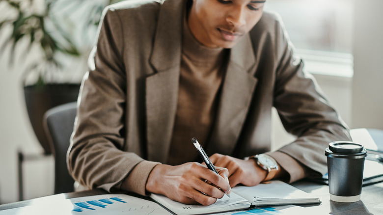 A man takes notes in a planner with coffee nearby.