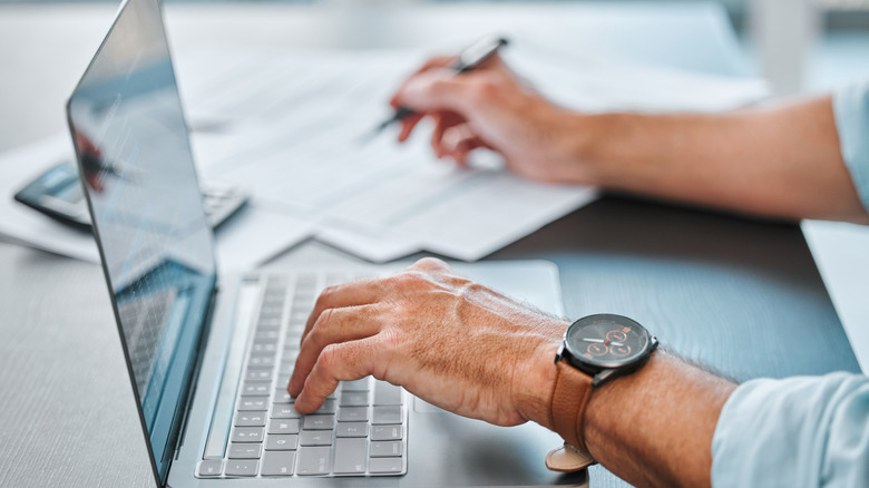 A man's hands are shown working on a computer while taking notes on paper.