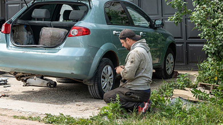 A man crouches to work on a car in a driveway.